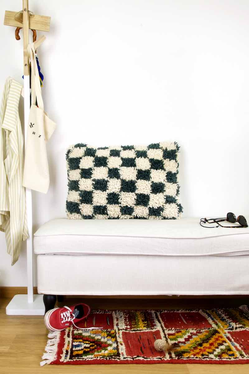 Checkered Moroccan Fluffikon throw pillow standing on a white bench. A red carpet is in front of the bench. The Berber pillow size is 40x60 cm.