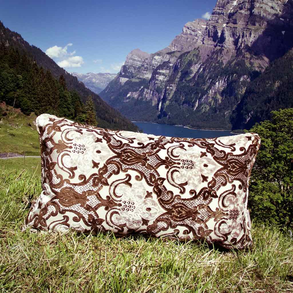 XXL Kissen in Braun vor Schweizer Bergen. Das Kissen steht auf einer Wiese vor einem See.