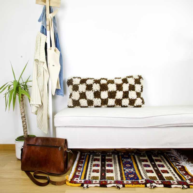 Brown white checkered Moroccan lumbar pillow on a white bench. Next to the bench is a brown leather bag. A Moroccan carpet is laying in front of the bench.