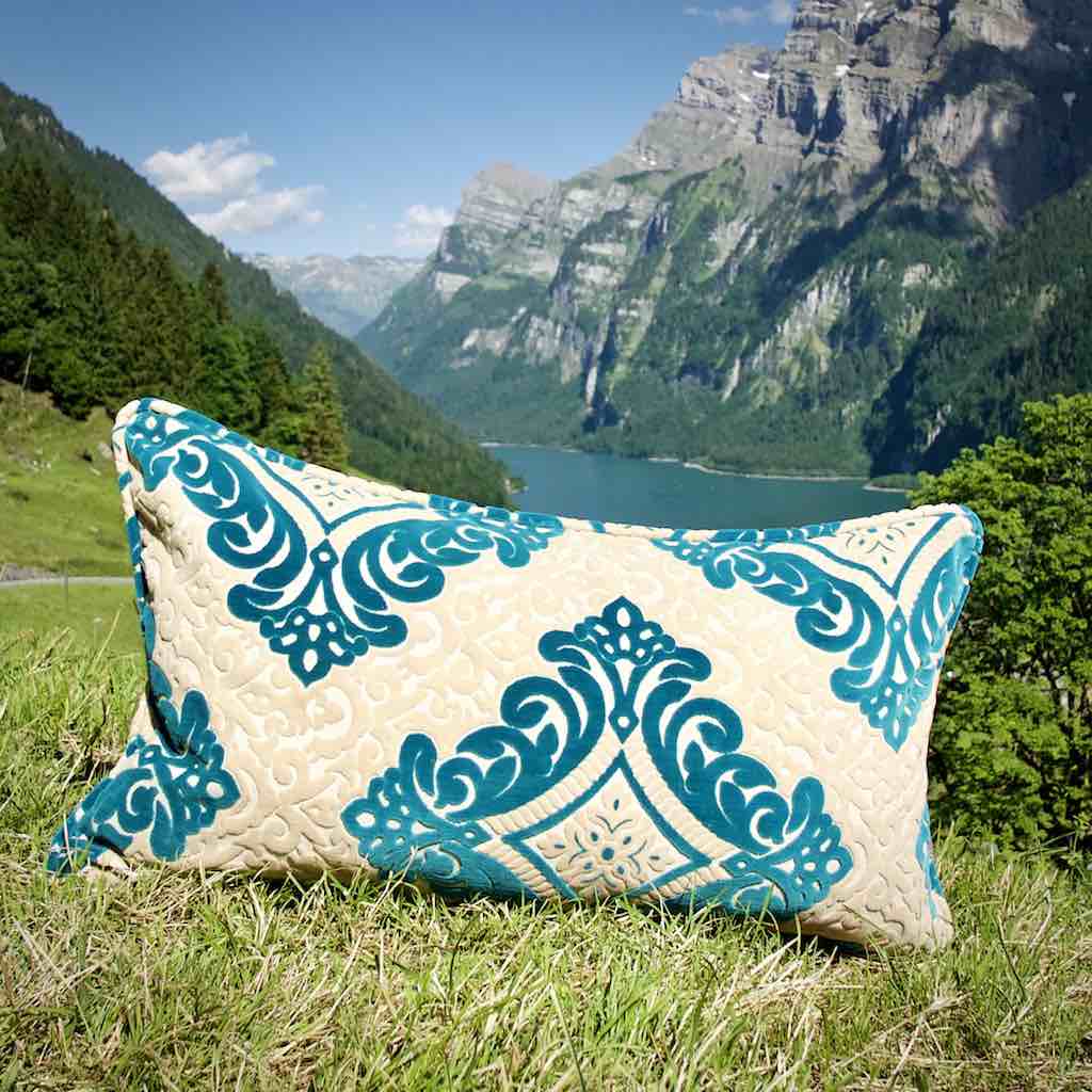 XXL Zierkissen vor Schweizer Berglandschaft. Das grosse Kissen steht auf einer Wiese vor einem blauem See.
