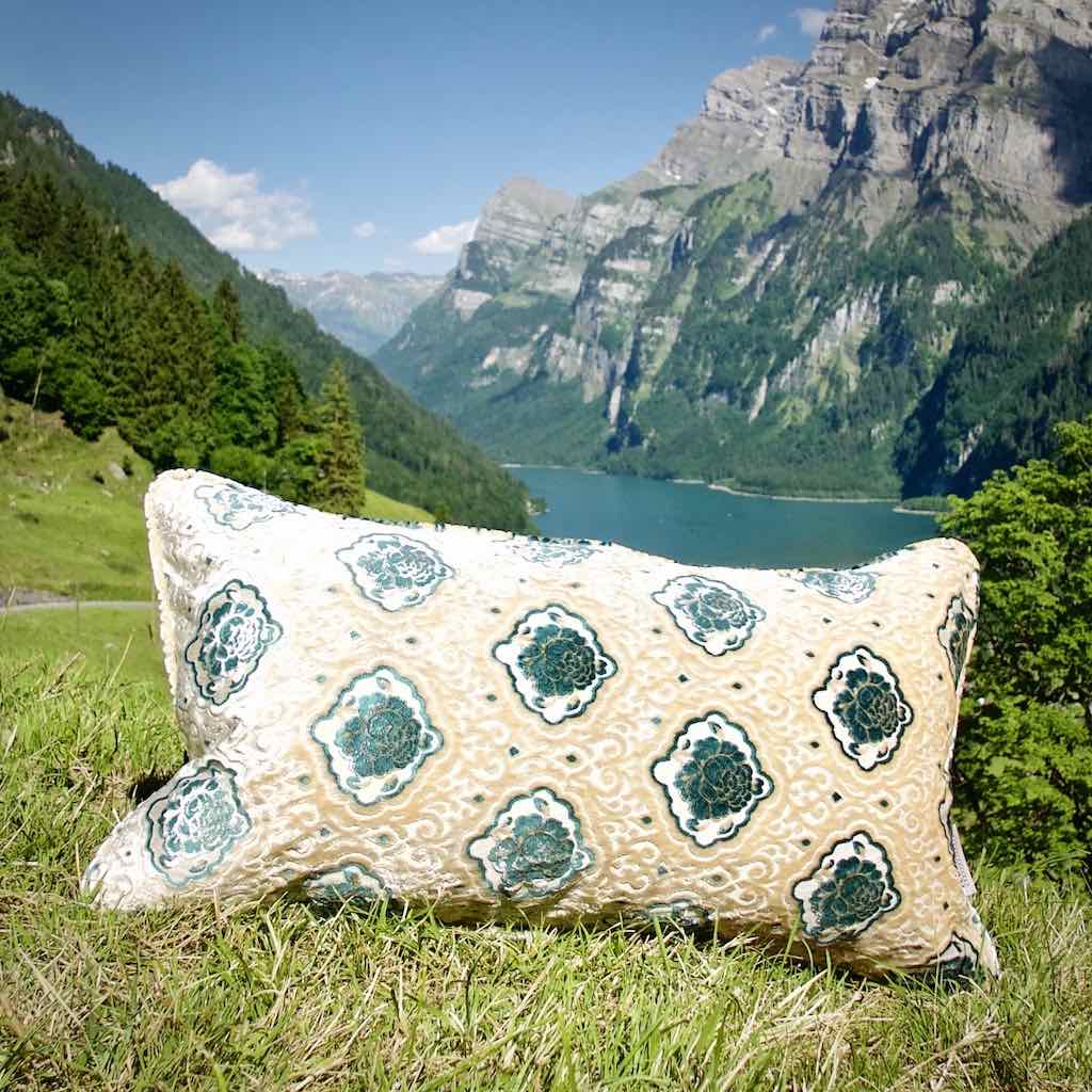 Grosses Fluffikon Sofakissen in den Schweizer Bergen. Das steht auf einer Wiese vor einem See.