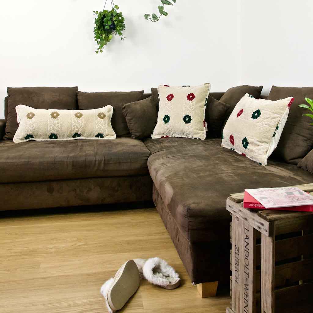 Three Fluffikon pillows on a brown sofa. The pillows have different sizes. A pair of slippers is in front of the couch.