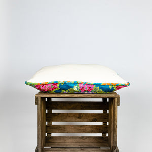 Blue Fluffikon decorative rectangular pillow made from shiny brocade with colourful flowers embroidery. The side of the pillow is shown. Pillow lies on a wooden box.