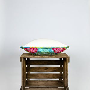 Blue Fluffikon decorative square pillow made from shiny brocade with colourful flowers embroidery. The side of the pillow is shown. Pillow lies on a wooden box.