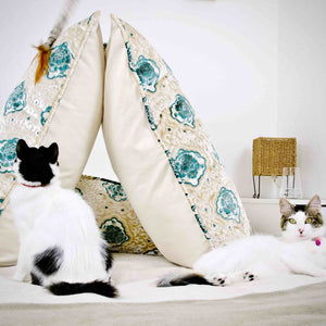 Two cats and two oversize Fluffikon throw pillows in a bed room. The cats are playing next to the pillows.