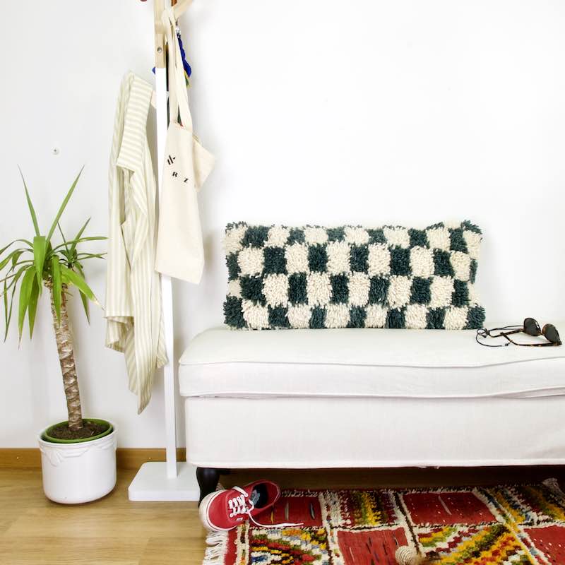 A Checkered Moroccan lumbar wool pillow on a white bench. The pillow is grey-blue white/beige checkered. 