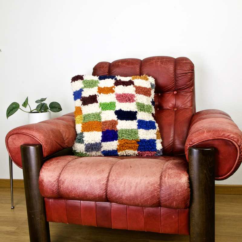 Colorful checkered Fluffikon throw pillow in the living room. The pillow is placed on a red leather chair.