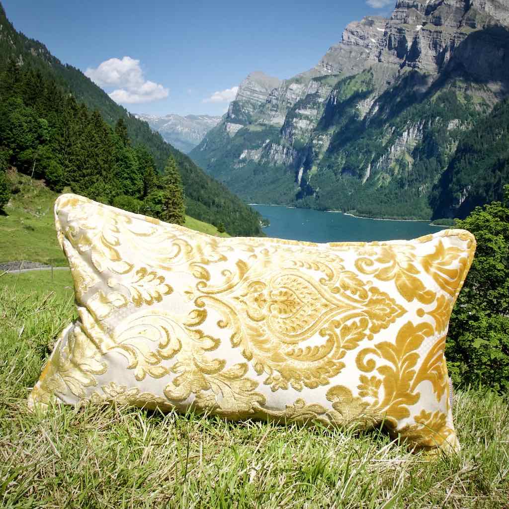 Grosses goldenes Kissen in den Schweizer Bergen. Das Kissen auf einer Wiese vor einem blauem See.