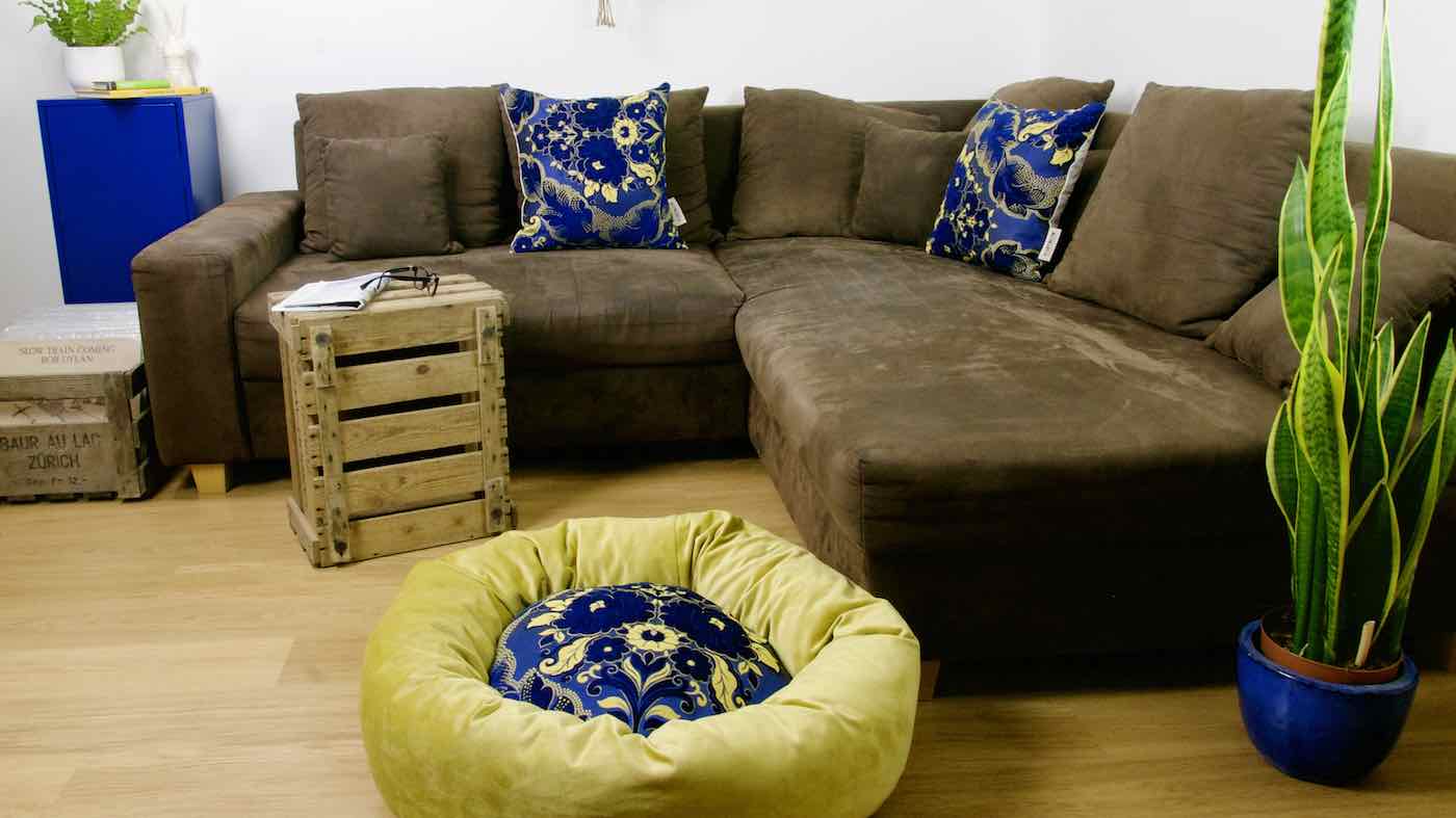 Blue golden Fluffikon dog bed in front of brown couch. The wool-filled dog is made from velvet fabrics.