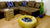 Blue golden luxury round dog bed in front of a brown couch. Two throw pillows on the couch with same pet bed material.