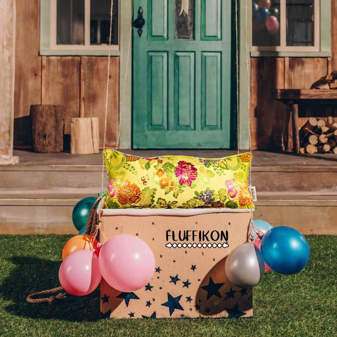 Yellow Fluffikon brocade pillow in a carton box with colourful balloons in front of a house.
