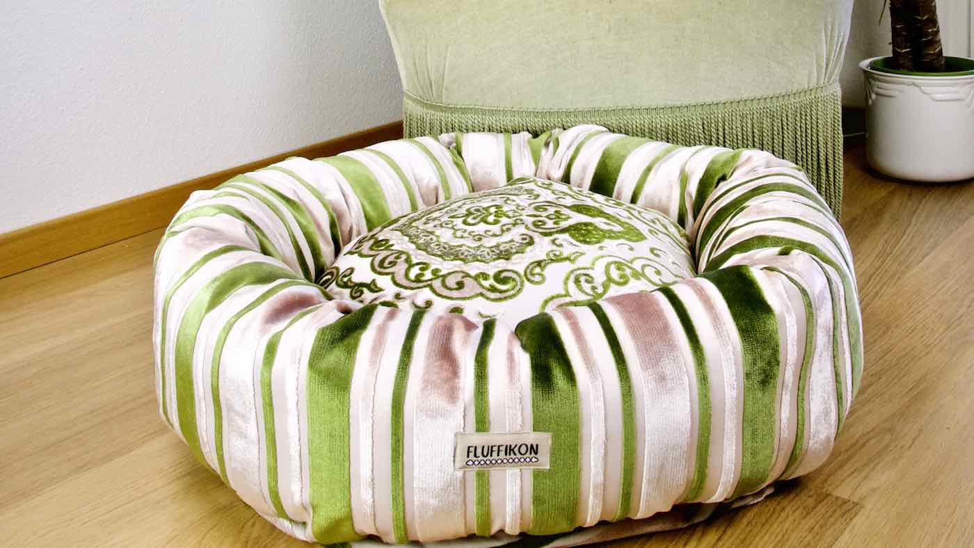 Grünes Fluffikon Hundebett vor einem grünen Sessel. Das edle Hundebett besteht aus grünem und beigen Samt.