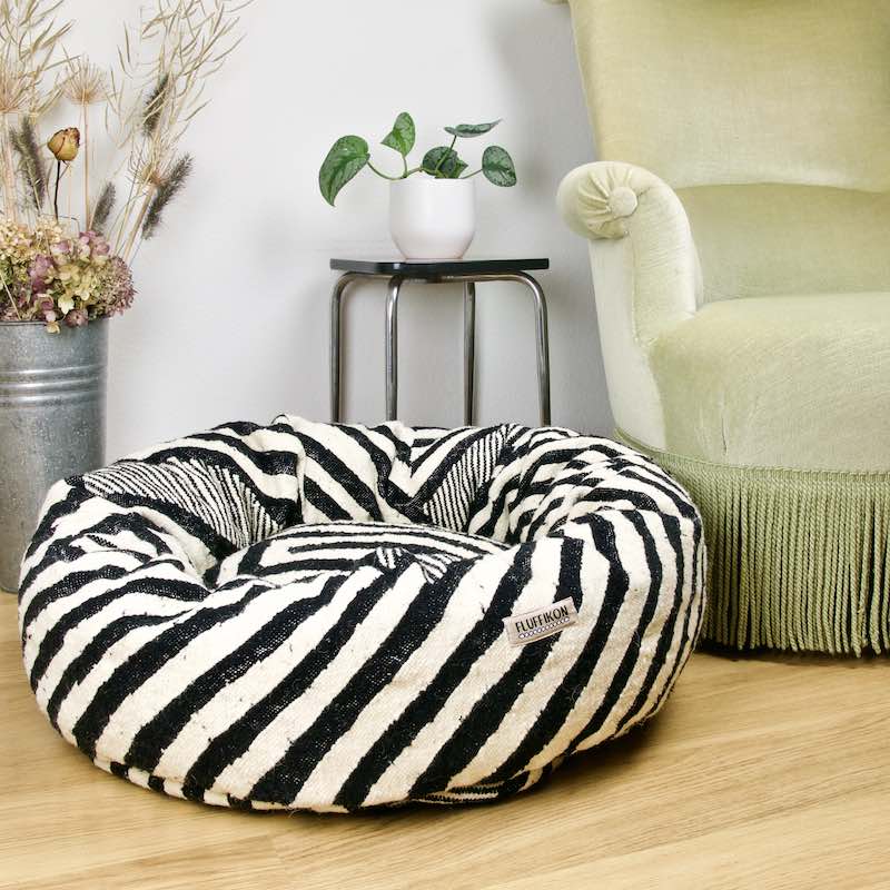 Schwarz weisses Donut Fluffikon Katzenbett, hergestellt aus einem Kelim Wollteppich. Das Bett steht vor einem grünen Sessel.