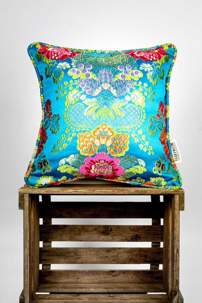 A square Fluffikon brocade pillow stnading on a wooden box.