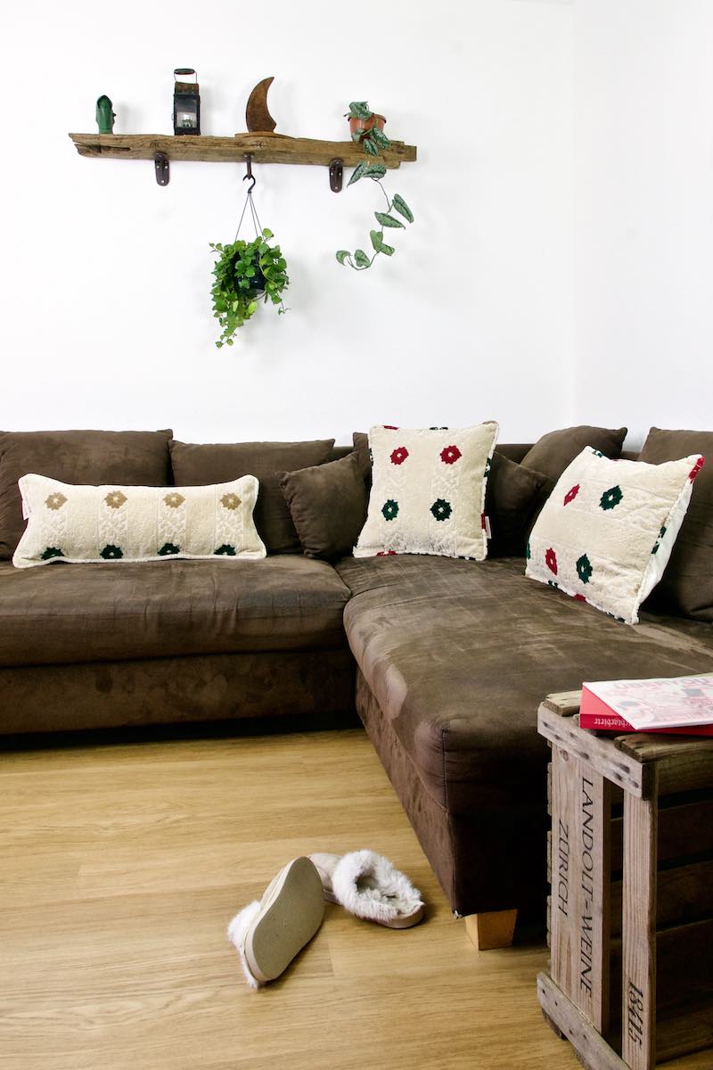 Three beige Fluffikon pillows on a brown sofa. The pillows have red and green flower patterns.