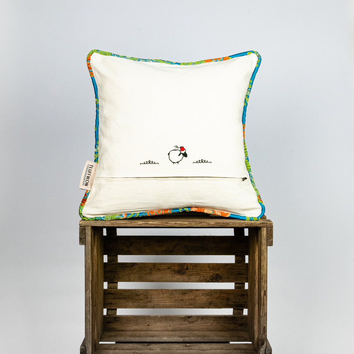 Blue decorative square pillow made from shiny brocade with colourful flowers embroidery. Back of the pillow is shown. Pillow stands on a wooden box.