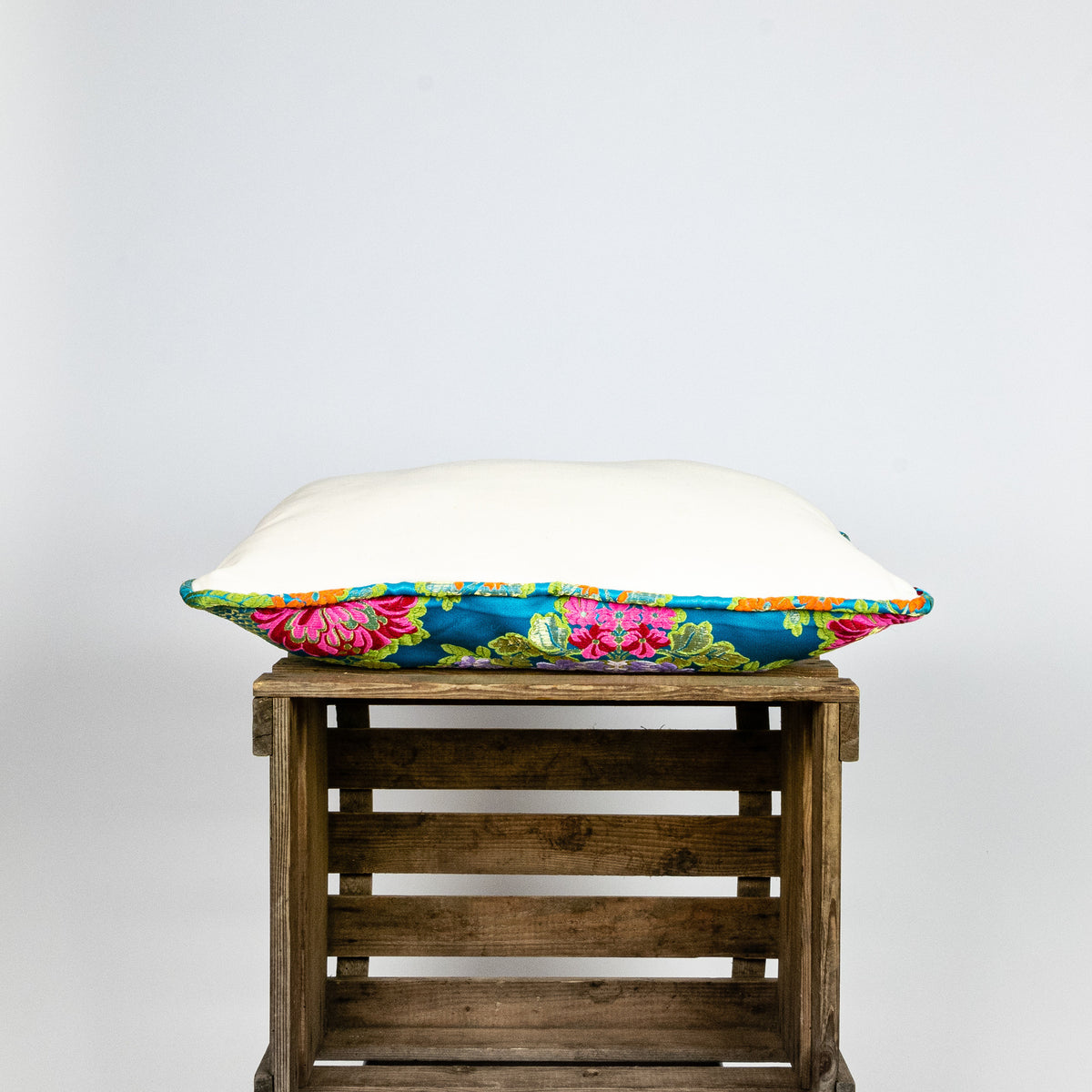 Blue Fluffikon decorative rectangular pillow made from shiny brocade with colourful flowers embroidery. The side of the pillow is shown. Pillow lies on a wooden box.