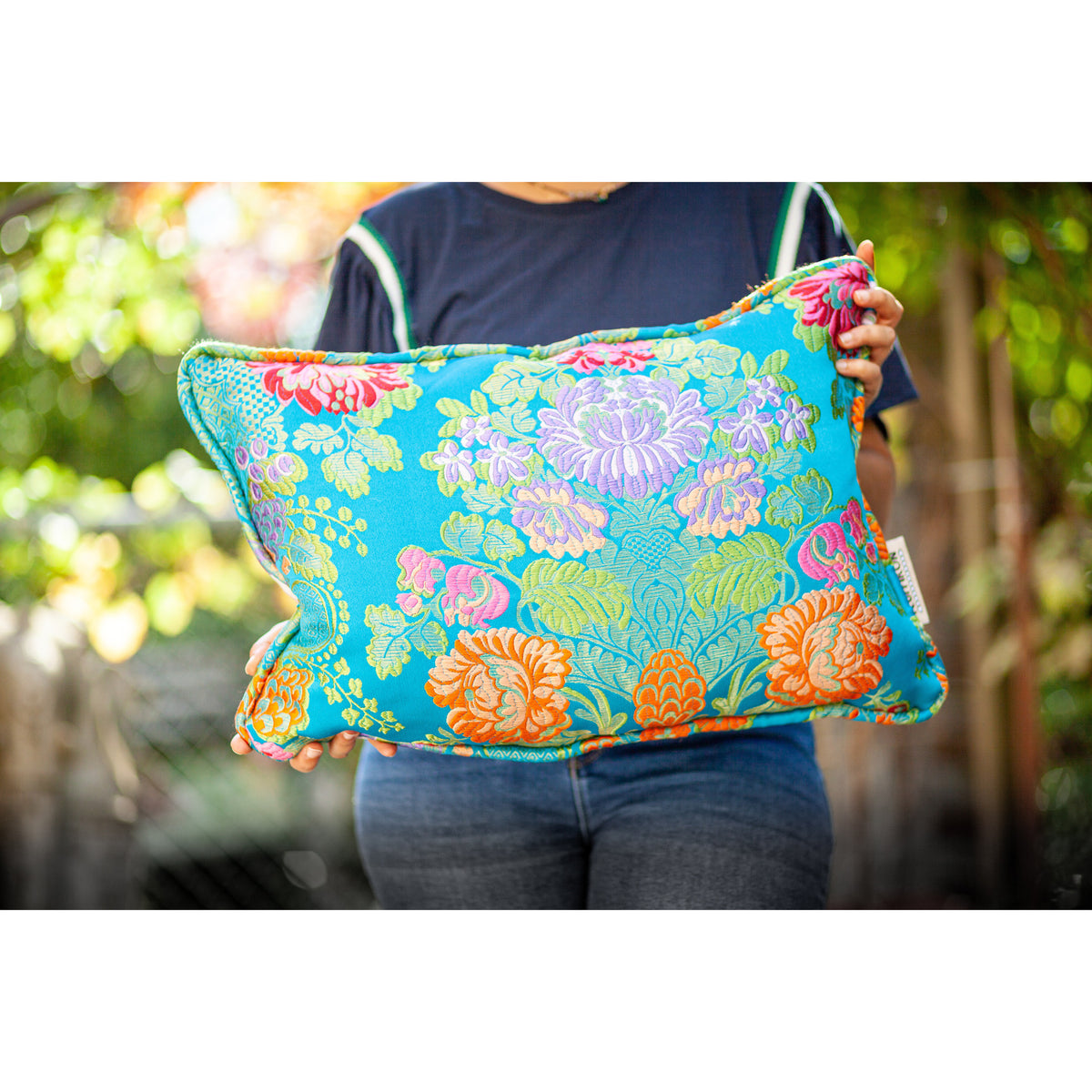 Woman holds blue Fluffikon decorative square pillow made from shiny brocade with colourful flowers embroidery.