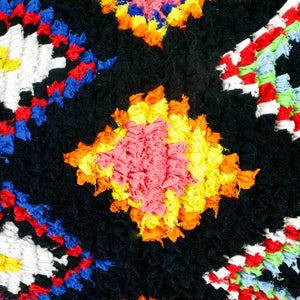 Close zoom on a black Fluffikon Boucherouite lumbar pillow. The Moroccan pillow is made from upcycled clothes. It has colorful patterns. A yellow red tile pattern is shown in detail.