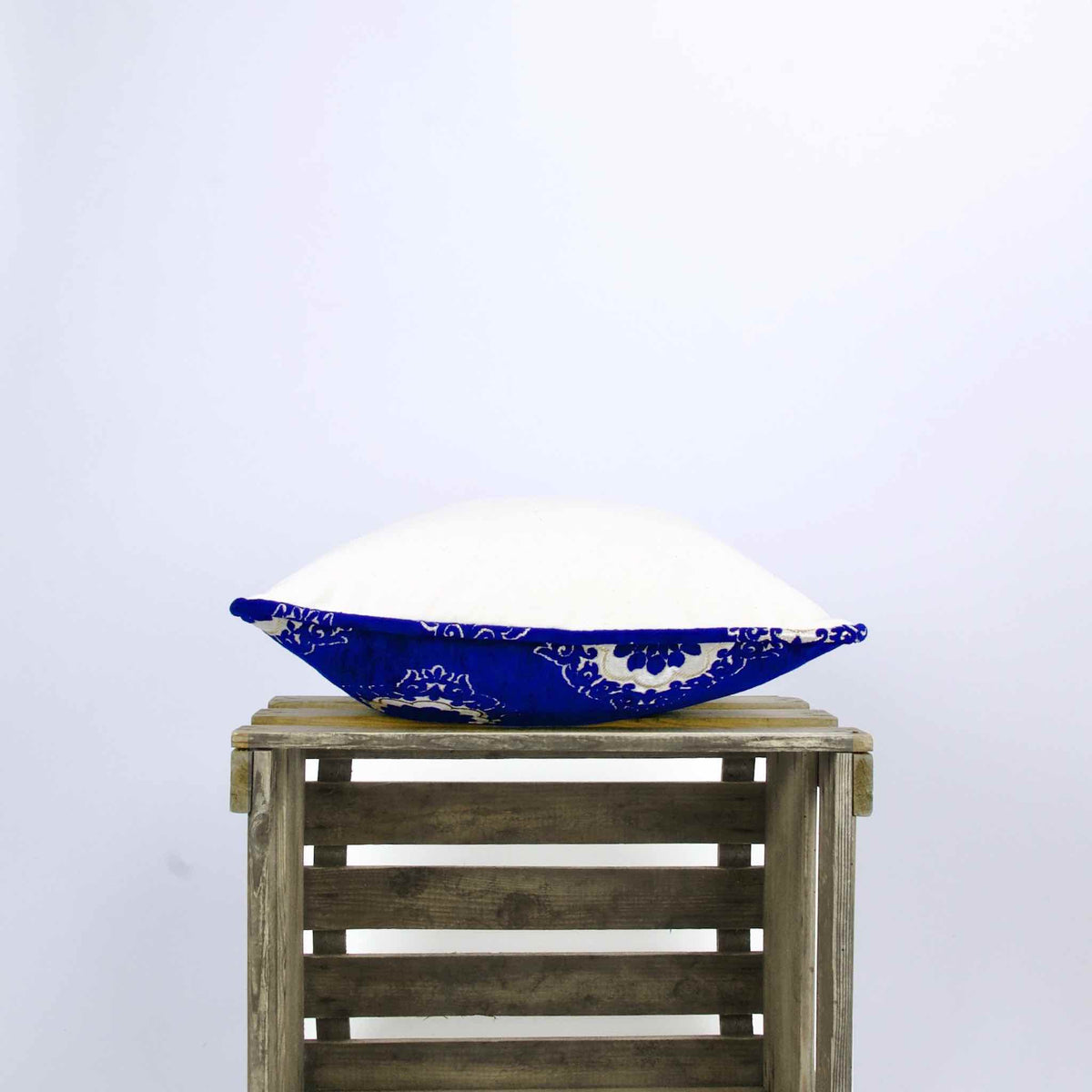 Side view of a Fluffikon sapphire blue velvet throw pillow. The pillows lies on a wooden box.