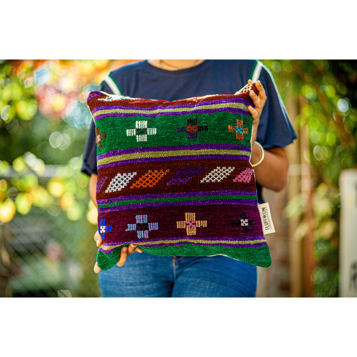 Fluffikon brown green decorative pillow with colourful berber symbols. It is made from a unique handmade thread rug held by a woman.